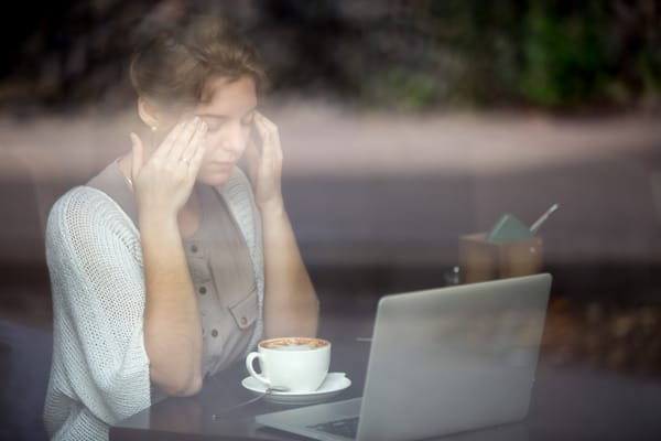 Stressed woman sitting at a café table with a laptop, holding her head from emotional exhaustion.
